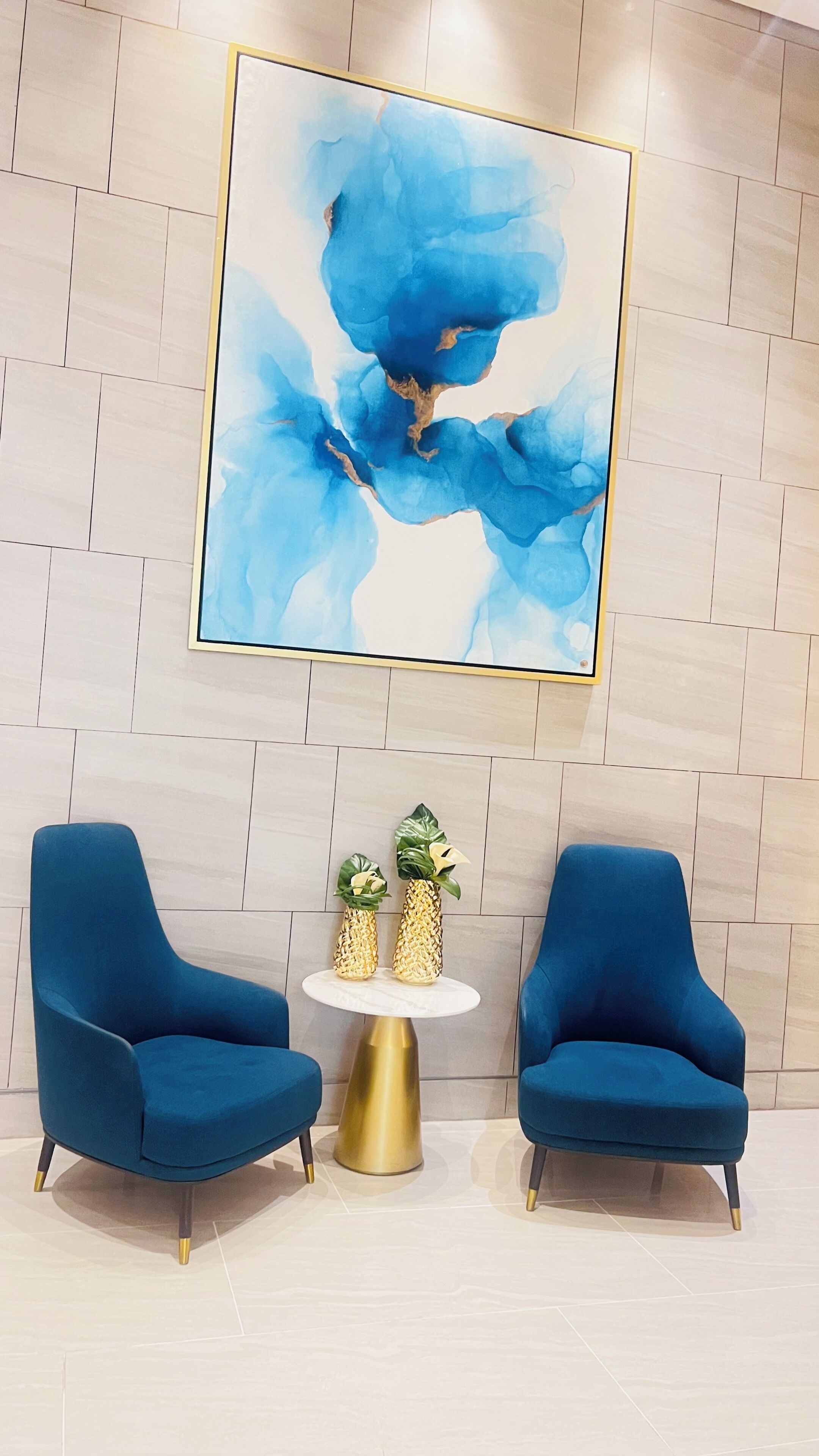 Una habitación con dos sillones azules, una mesa redonda dorada con dos jarrones decorativos, y un cuadro abstracto azul grande en la pared.