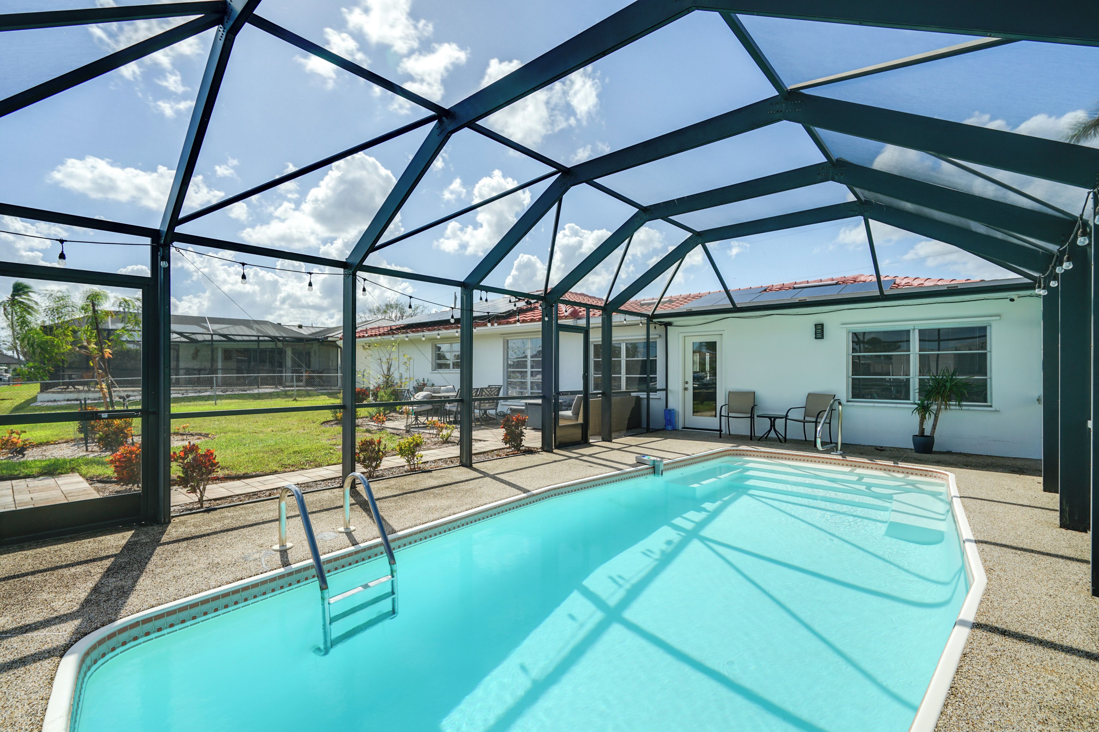 A covered swimming pool with a metal frame, a patio area with chairs, and a view of a house with a red-tiled roof.