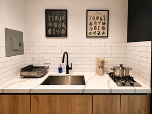 Una cocina moderna con salpicadero de azulejos blancos, un grifo negro, un lavamanos de acero inoxidable, una estufa a gas y dos ilustraciones enmarcadas de un jardín de hierbas y vegetales en la pared.