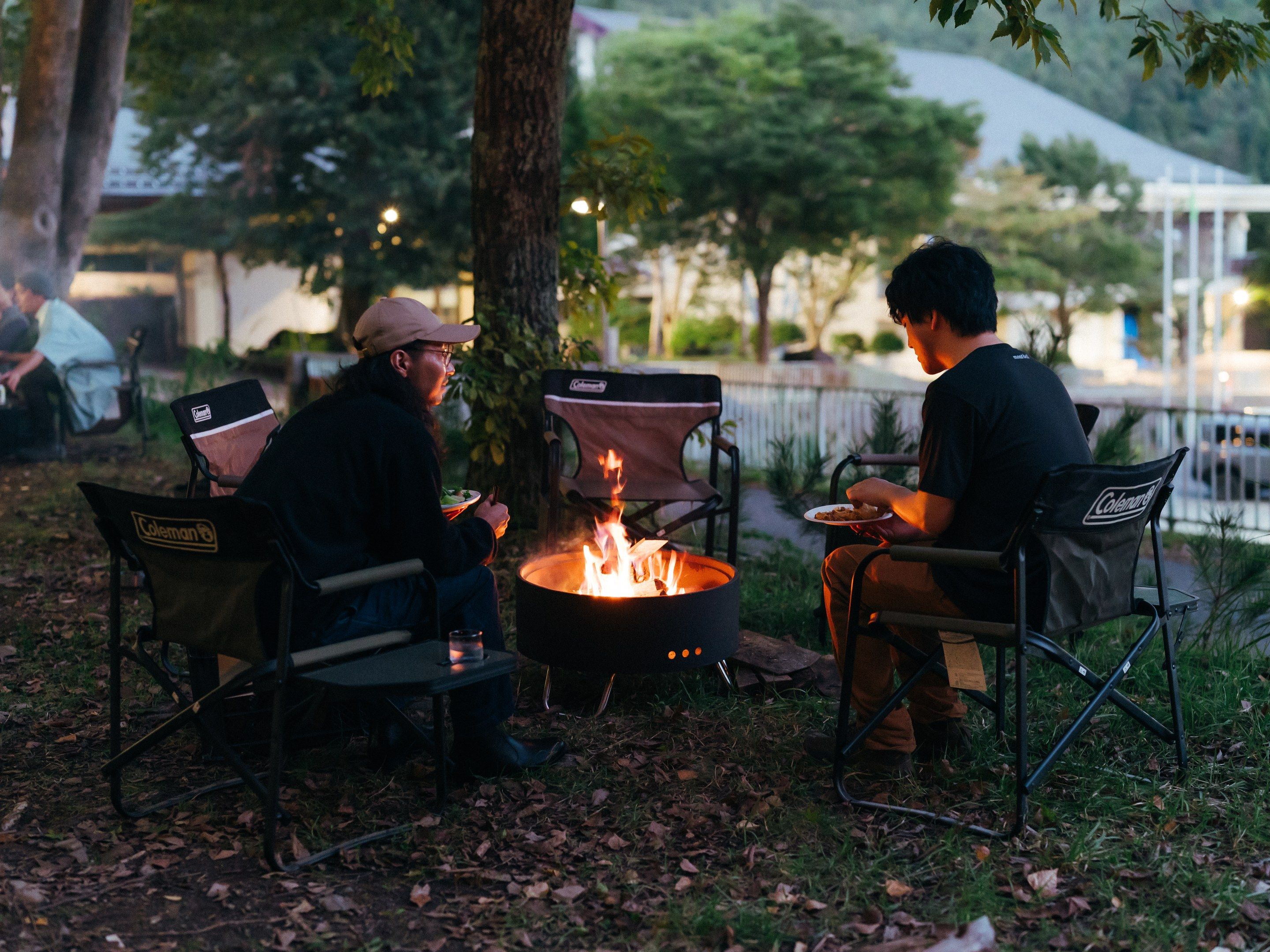 Two people seated at a campfire with camping chairs, surrounded by trees and a body of water in the background.