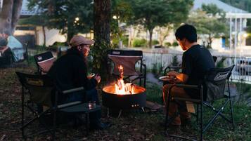 Two people seated at a campfire with camping chairs, surrounded by trees and a body of water in the background.