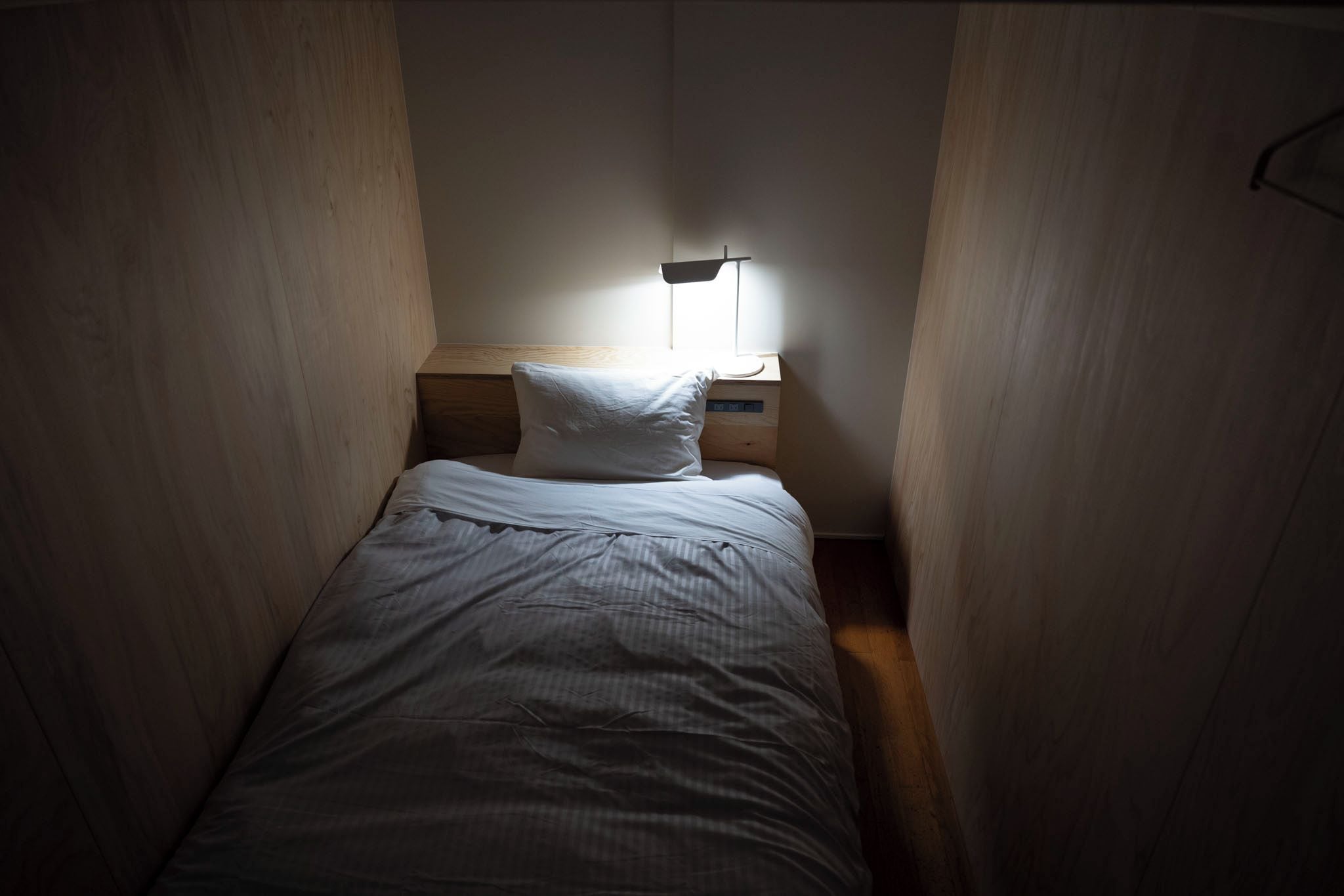 Una habitación pequeña con una cama individual, una mesita de noche y una lámpara en la pared.