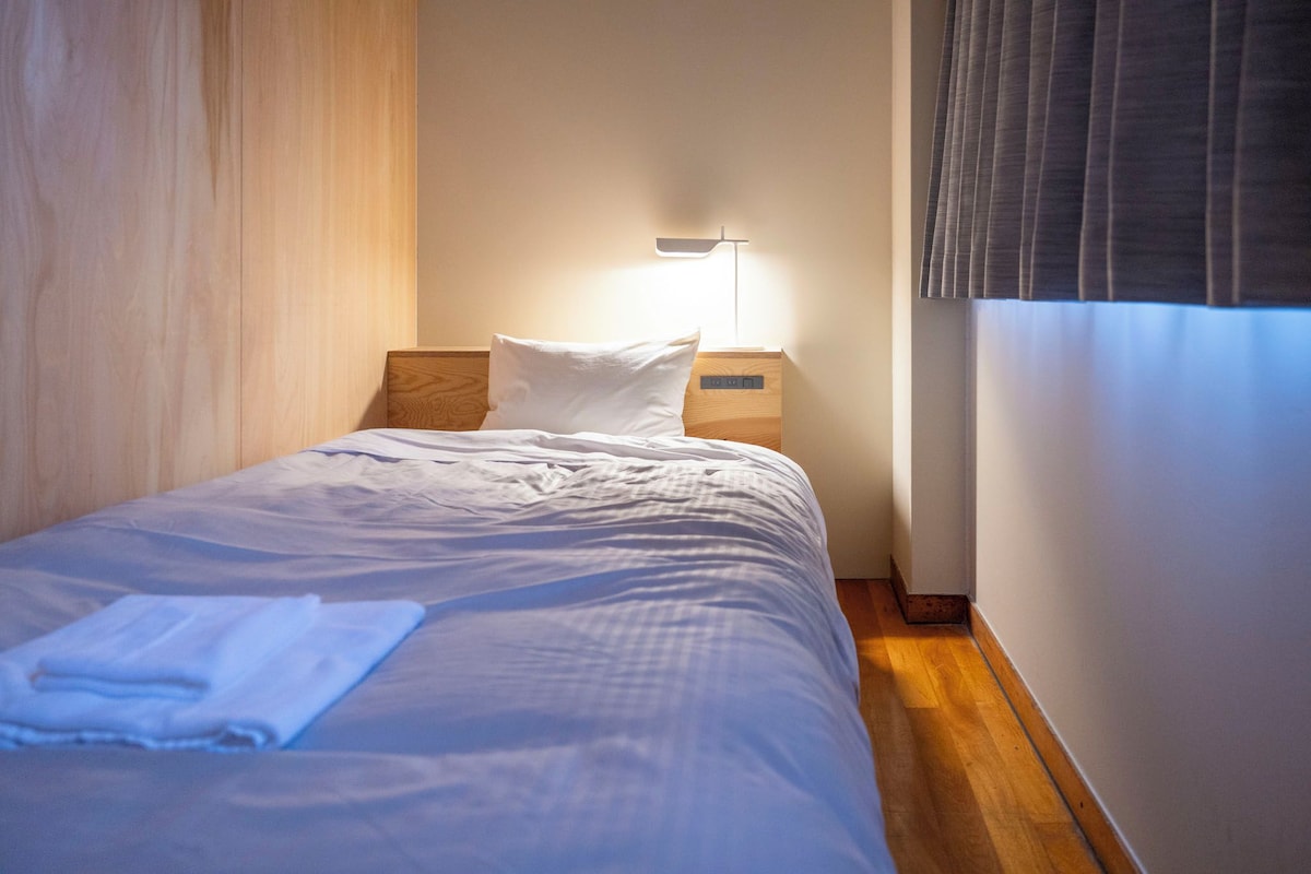 A neatly made bed with white linens and a folded towel, a bedside table with a lamp, and a window with blinds.