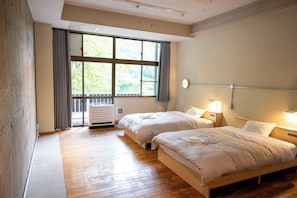 A hotel room with two beds, a large window with curtains, a radiator, and a bedside table with a lamp.