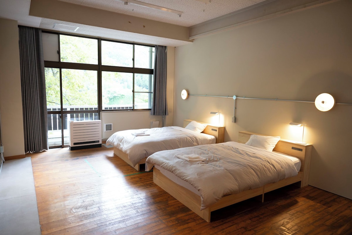 A hotel room with two beds, large windows, a radiator, and wall-mounted lights.