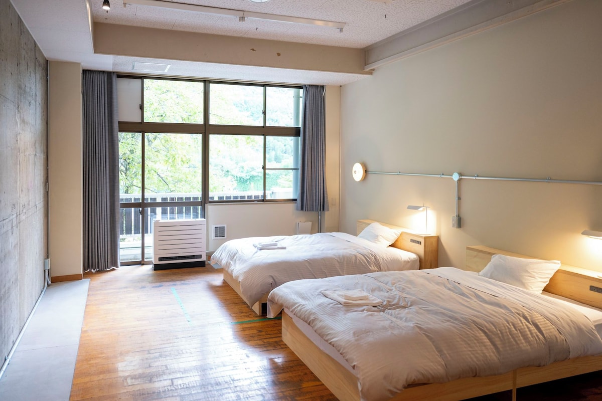 A hotel room with two beds, a large window with curtains, a ceiling light, and a wall-mounted heater.