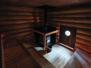 Interior de una cabaña de madera con un estufa de leña en el centro, una ventana y una puerta.