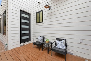 A covered outdoor seating area with two chairs and a small table, a black door, and a window on a white siding house.
