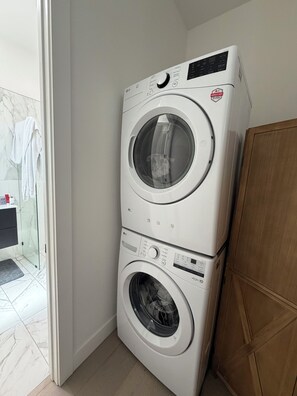 A stacked washer and dryer unit in a corner of a room with a glass shower door and a wooden cabinet.