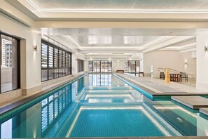 A long indoor swimming pool with clear water, surrounded by a modern interior with large windows and a high ceiling. - 1BR City Retreat with Pool & Gym Zodiak Stays (Auckland)