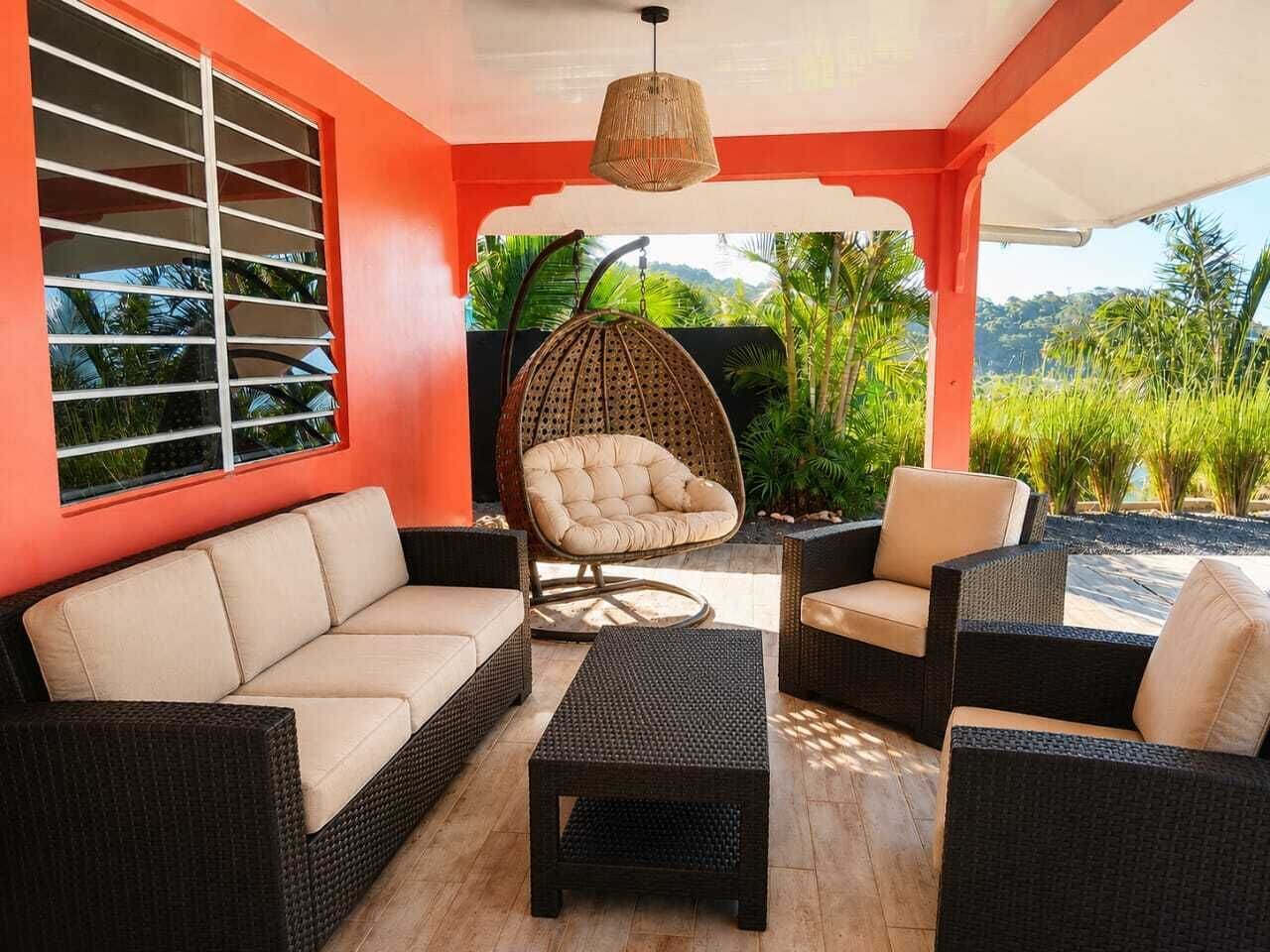 A covered outdoor seating area with wicker furniture, a cushioned chair, and a view of greenery.
