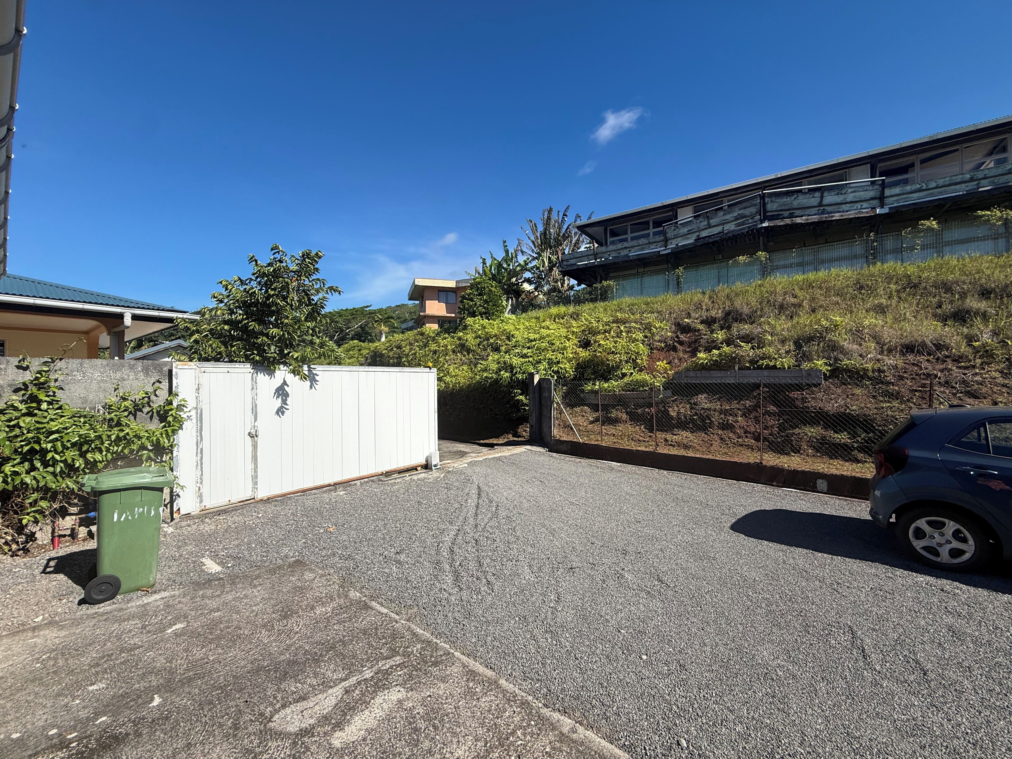 A parking area with a car, a white gate, and a green trash bin.