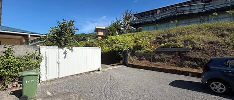 A parking area with a car, a white gate, and a green trash bin.