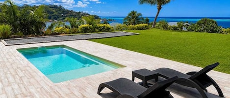 A rectangular swimming pool with a wooden deck, two lounge chairs, and a view of the ocean and hills.