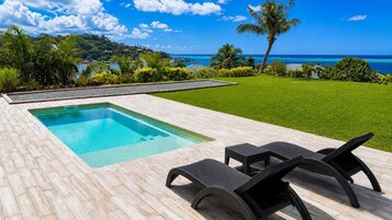 A rectangular swimming pool with a wooden deck, two lounge chairs, and a view of the ocean and hills.