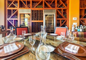 Una mesa de comedor preparada para cuatro, con copas de vino, platos y cubiertos, rodeada por una bodega con varias botellas.
