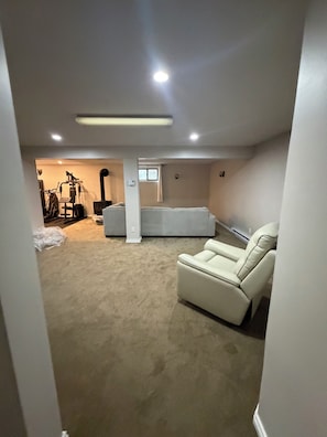 A carpeted basement with a white leather recliner, a workout machine, and a window.