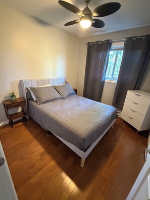 A bedroom with a bed, a nightstand, a dresser, a ceiling fan, and a window with curtains.