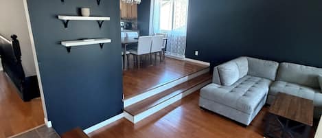 Uma sala de estar moderna com um sofá seccional cinza, uma área de jantar com mesa e cadeiras, e uma cozinha ao fundo.