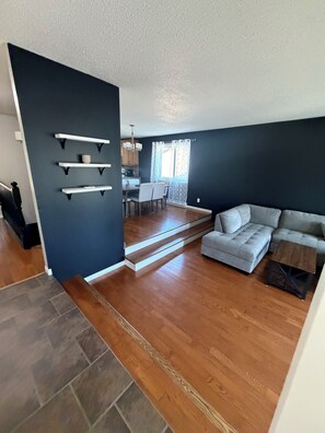 A modern living room with a grey sectional sofa, a dining area with a table and chairs, and a kitchen in the background.