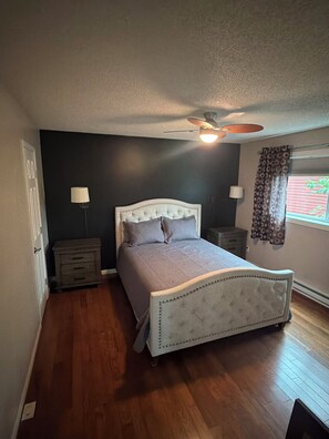 Un dormitorio con una cama individual, un ventilador de techo, dos mesitas de noche y una ventana con cortinas.