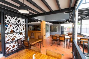 Un'area ristorazione interna con tavoli e sedie in legno, soffitto nero con travi a vista e un pannello decorativo bianco.