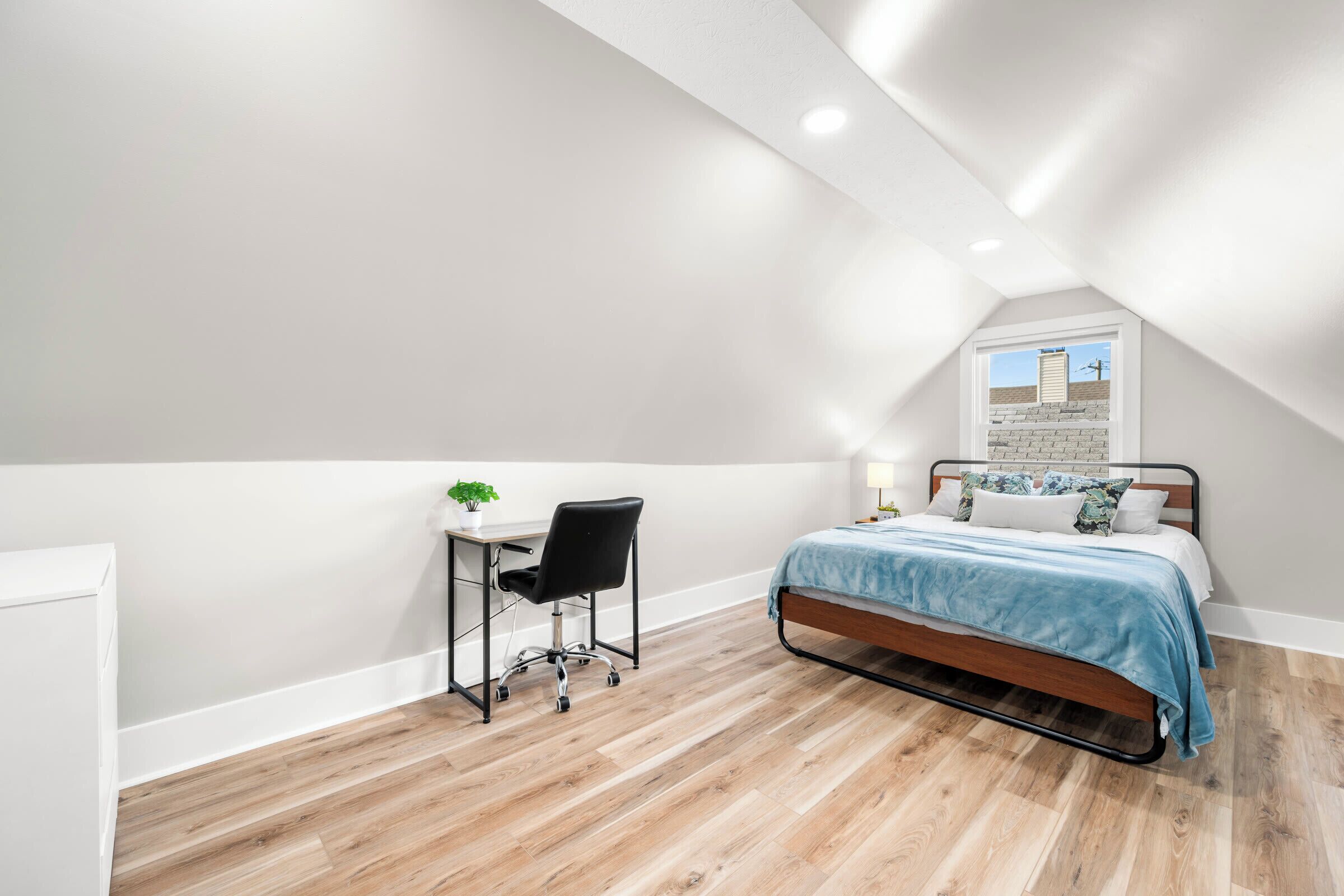 A bedroom with a sloped ceiling, a bed with a blue bedspread, a desk with a chair, and a window with a view of a building.