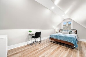 Ein Schlafzimmer mit schrägen Wänden, einem Bett mit blauer Bettdecke, einem Schreibtisch mit Stuhl und einem Fenster mit Blick auf ein Gebäude.