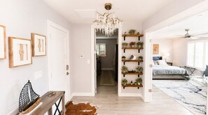Un interior moderno con candelabro, estanterías de madera con plantas, cama con vistas y un pasillo que lleva a otra habitación.