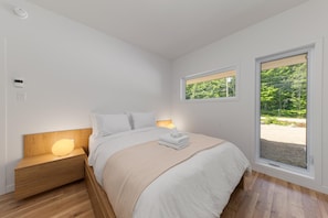Een slaapkamer met een bed, bedlampen, een raam met uitzicht op groen en een houten nachtkastje.