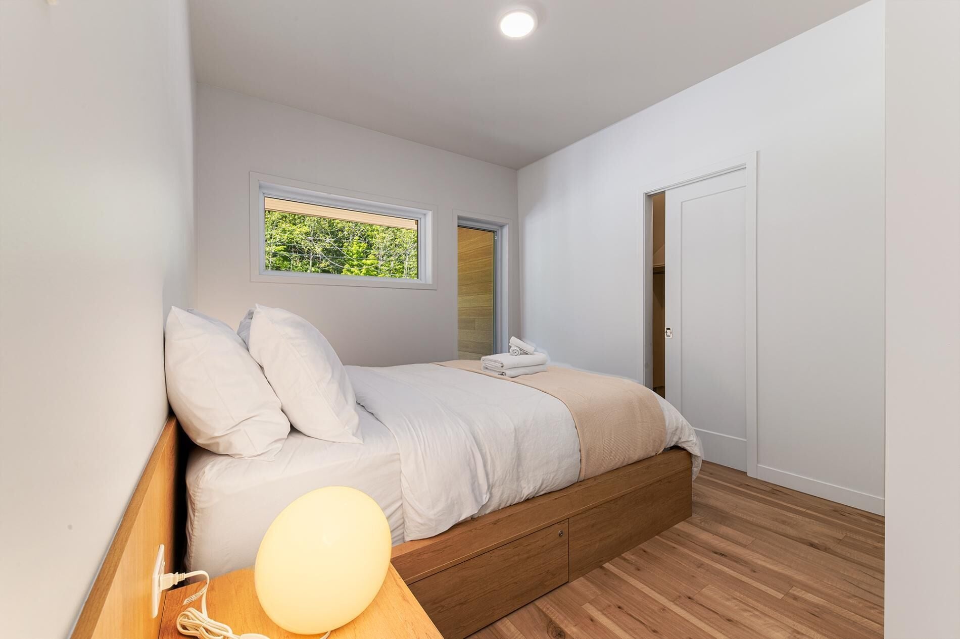 A bedroom with a bed, a bedside lamp, a window with a view, and a door.