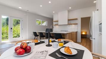 A modern kitchen with white cabinets, a breakfast table with apples, croissants, and a newspaper, and a view of greenery outside.