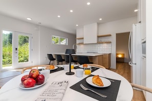 Una cocina moderna con armarios blancos, una mesa de desayuno con manzanas, cruasanes y un periódico, y vistas al exterior con vegetación.