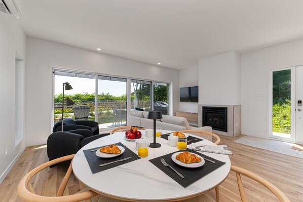 A modern living room with a dining area, large windows, a fireplace, and a television.