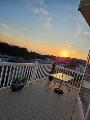A wooden deck with a white railing, a table, and chairs, overlooking a residential area during sunset.
