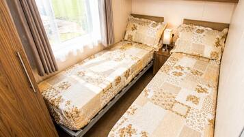 A compact room with two single beds, a wooden wardrobe, a small table with a lamp, and a window with curtains.