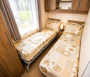 A compact room with two single beds, a wooden wardrobe, a small table with a lamp, and a window with curtains.