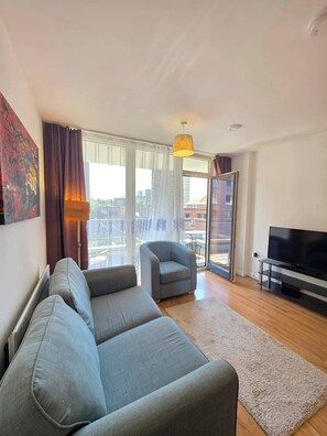 Ein modernes Wohnzimmer mit Sofa, Sessel, Fernseher und einem großen Fenster mit Vorhängen.