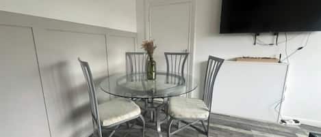 Un comedor con una mesa redonda de vidrio, cuatro sillas, un florero con flores y un televisor de pantalla plana montado en la pared.