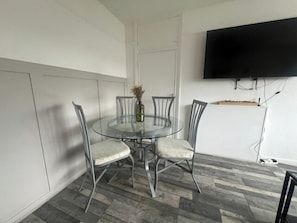 Un coin repas comprenant une table ronde en verre, quatre chaises, un vase avec des fleurs et un téléviseur à écran plat fixé au mur.