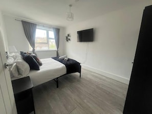 Une chambre à coucher avec un lit, une fenêtre avec des rideaux, un téléviseur à écran plat et un plancher en bois.