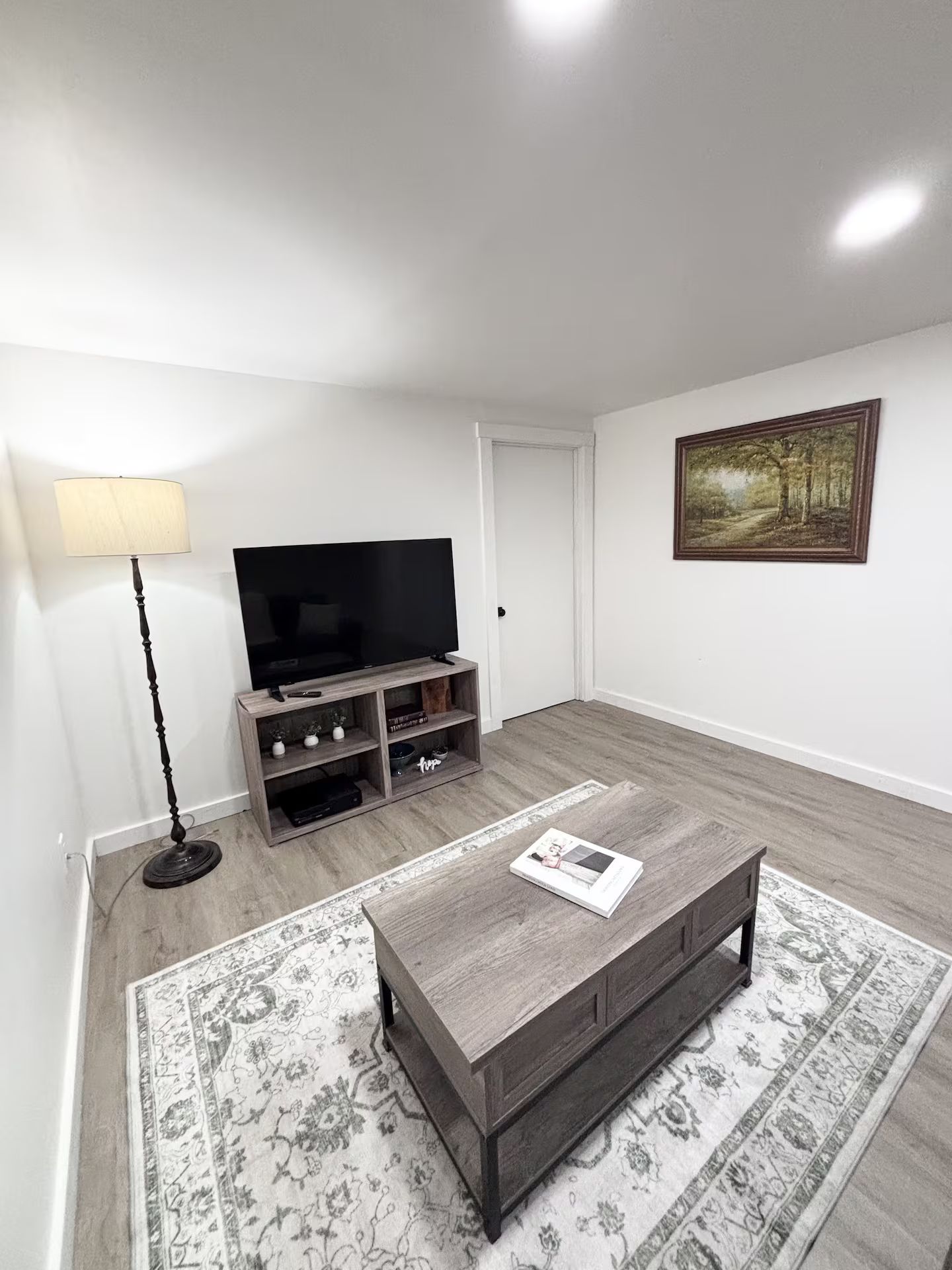 Una sala de estar con un televisor de pantalla plana sobre un soporte de madera, una lámpara de pie, una mesa de centro con una revista y un cuadro en la pared.