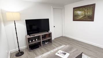 Una sala de estar con un televisor de pantalla plana sobre un soporte de madera, una lámpara de pie, una mesa de centro con una revista y un cuadro en la pared.