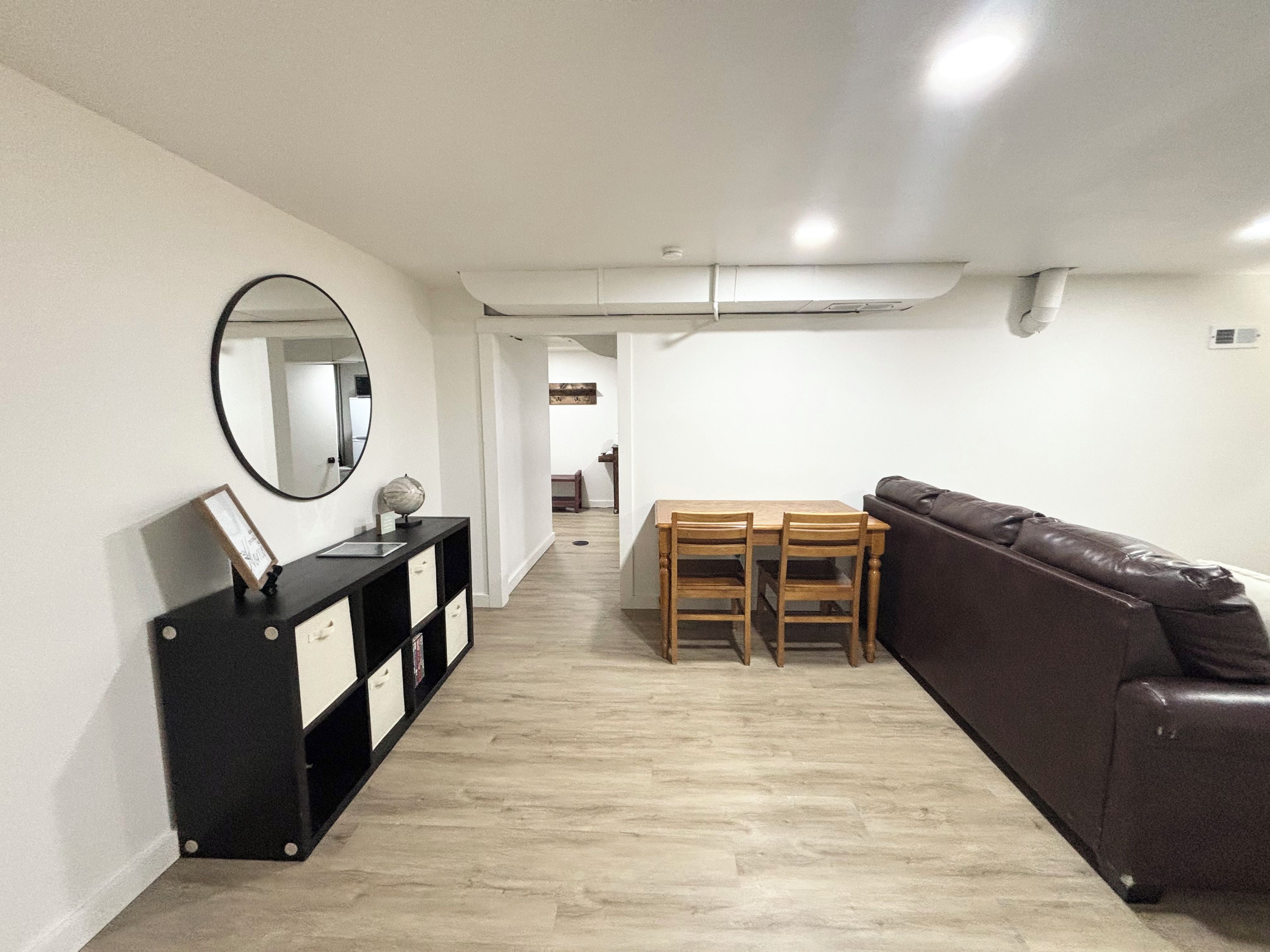 Una sala moderna con un sofá de cuero marrón, una mesa de comedor de madera con sillas, un mueble bajo negro con cajones y un espejo redondo en la pared.