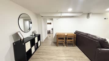 Una sala moderna con un sofá de cuero marrón, una mesa de comedor de madera con sillas, un mueble bajo negro con cajones y un espejo redondo en la pared.