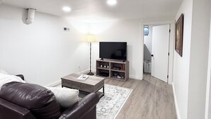 A modern living room with a leather sofa, a coffee table, a TV on a wooden stand, and a hallway leading to another room. - Spacious & Cozy Suite Getaway in KC Area · No Stairs (Blue Springs)