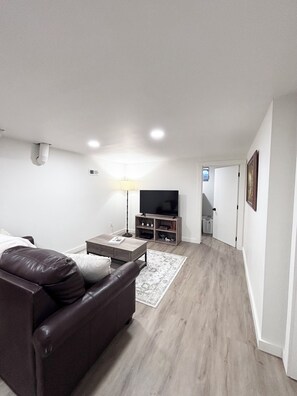 Un salon moderne avec un canapé en cuir, une table basse, un téléviseur posé sur un meuble en bois, et un couloir menant à une autre pièce.