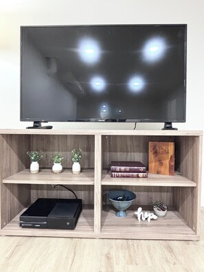 Un televisor sobre un soporte de madera con tres estantes debajo. El estante superior tiene dos jarrones blancos con plantas, el estante del medio tiene una pila de libros, y el estante inferior tiene un reproductor de DVD, un cuenco decorativo y una planta pequeña.