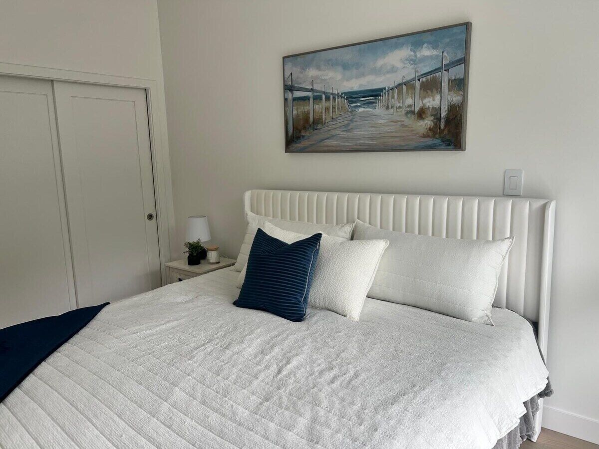 A bedroom with a white bed, a headboard, and a painting of a coastal scene.