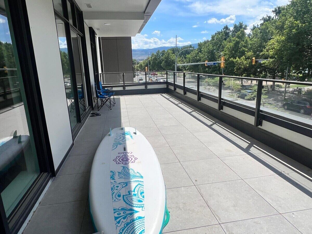 Un balcón con una tabla de surf, baranda de vidrio y vista a una carretera y árboles.